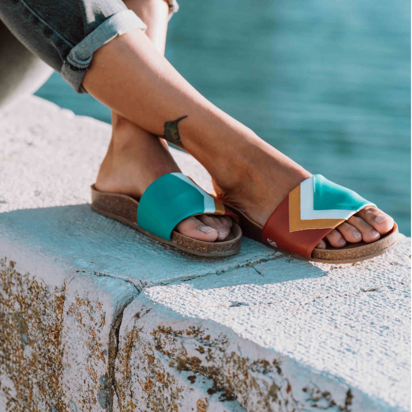 Mit PETA Vegan Award 2023 ausgezeichnete türkis-braune Sandalen; vegan mit Fußbett; Farben Ocean, Weiß, Senf, Terracotta; Marke Fünve; Modell Sunset; Mood auf Mauer