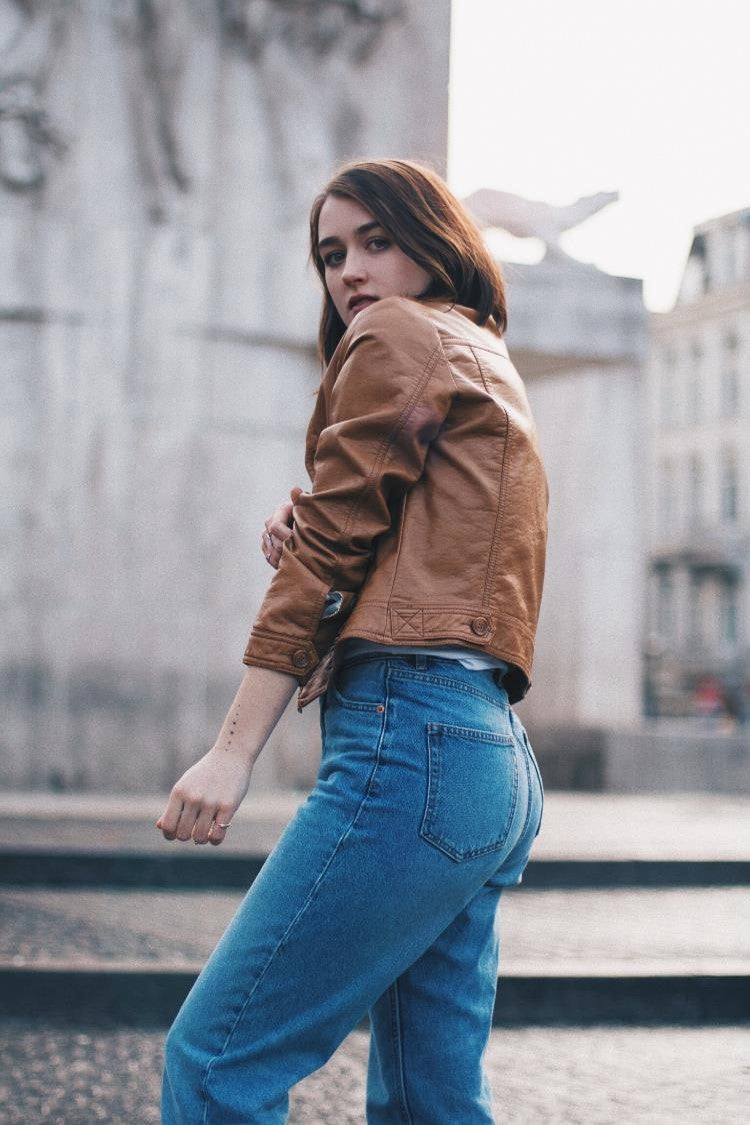 Fünve - eine junge Frau in brauner Kunstlederjacke und blauen Jeans blickt sich über die Schulter um